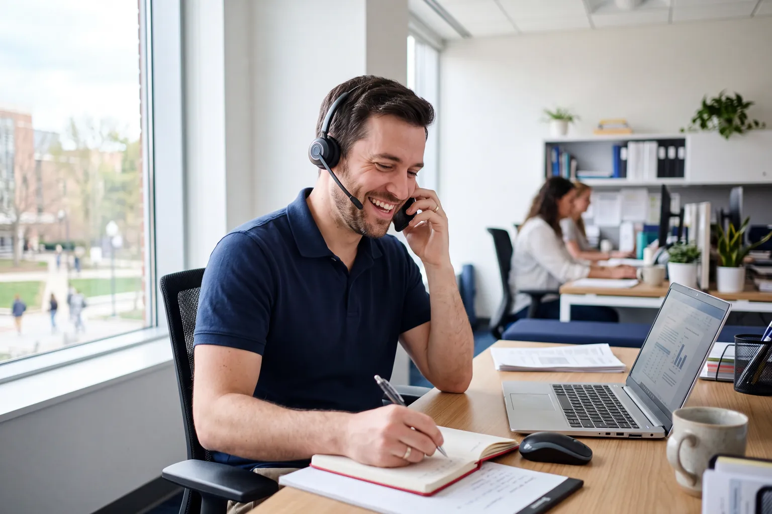 Clinical research coordinator in a navy polo smiling on a phone call and writing notes at his desk